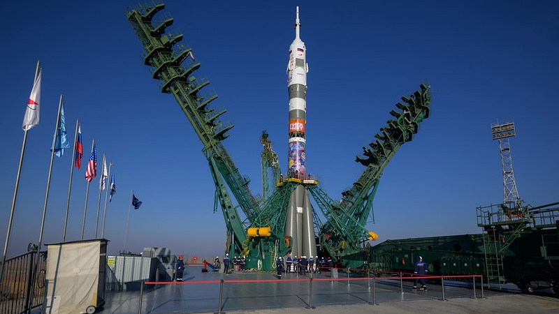 photo of a white rocket with a brown base standing at a launch pad beneath a blue sky