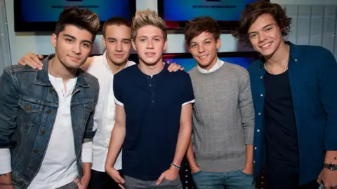 PA Media (left to right) Zayn Malik wears a denim jacket over a white t-shirt. He is stood beside Liam Payne, Niall Horan, Louis Tomlinson and Harry Styles. Liam is wearing a white shirt, Niall is wearing a navy t-shirt, Louis is wearing a grey jumper, and Harry is wearing a blue shirt over a navy t-shirt. 
