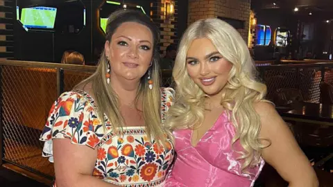 Breege Gormley Breege is wearing a white and multi-coloured dress with multi-coloured earrings. She has long dark brown hair and is standing beside her daughter, Jenny.  Her daughter has long curly blonde hair and is wearing a pink dress. They are both standing in a bar. 