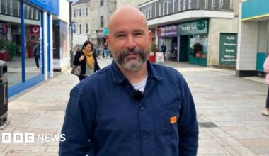 The two buildings which make up Trago Mills in Falmouth. One is wooden two-storey building and the other is a three-storey building along Arwenack Street. There is a hatched area in front of the wooden building and it has the nations' flags flying.