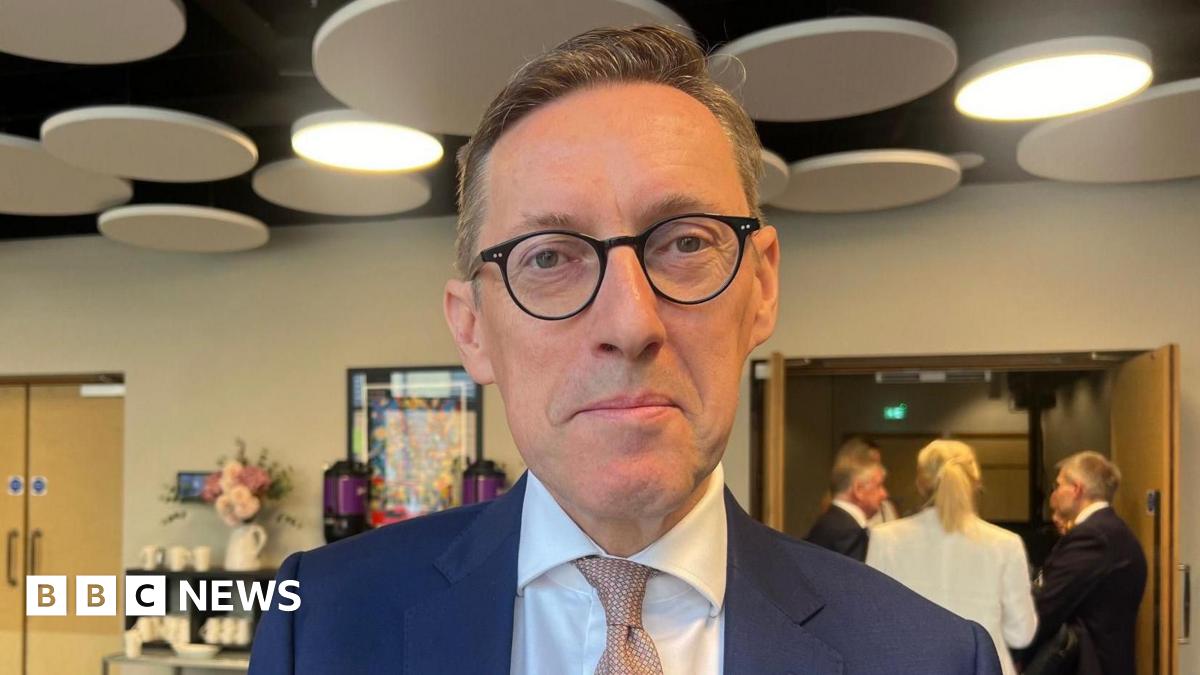 Deputy Ian Gorst is wearing a blue suit and white shirt with a pink tie.