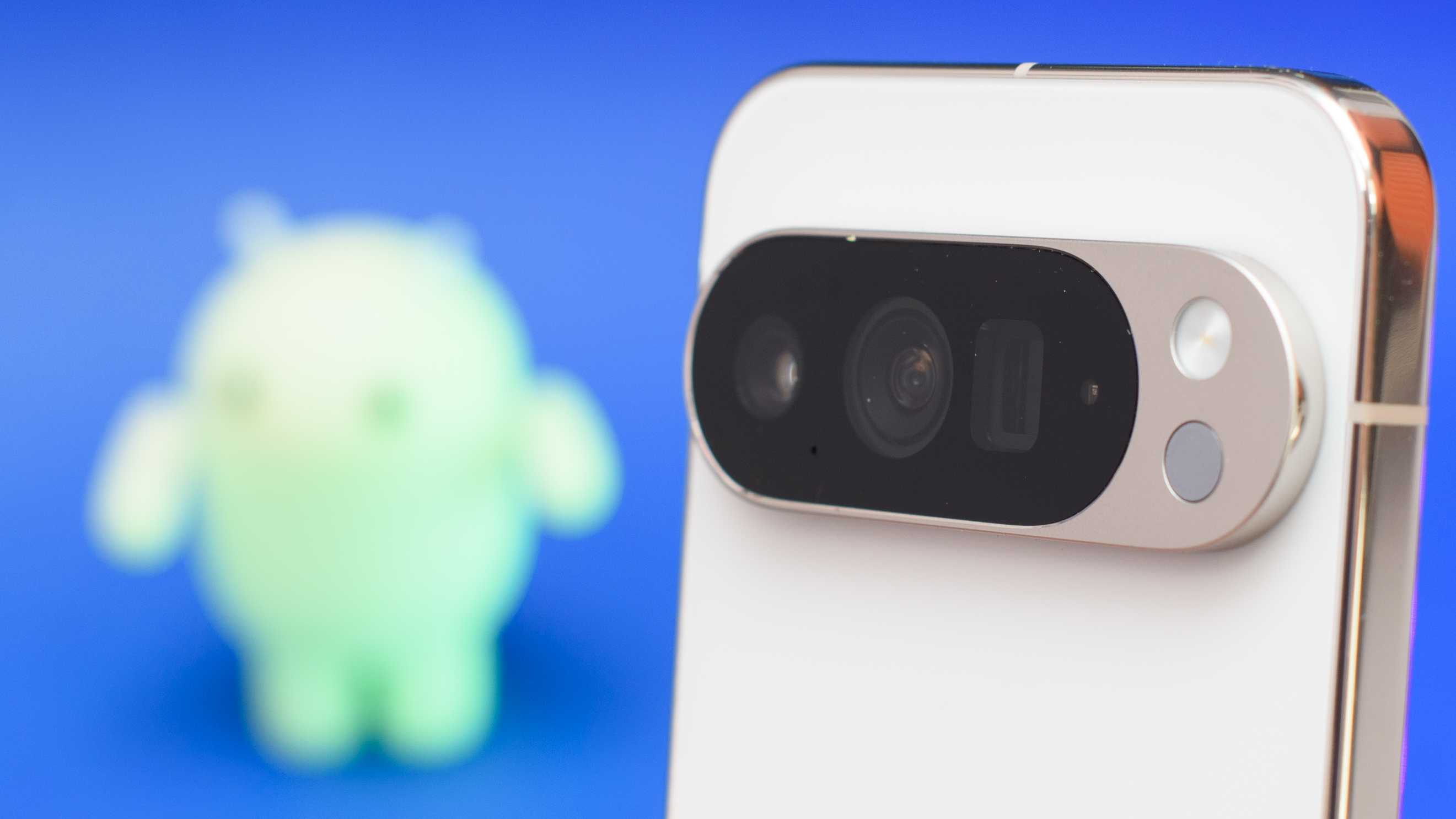 Google Pixel 10 Pro XL in Porcelain against a blue background with a green neon Android figurine in the background