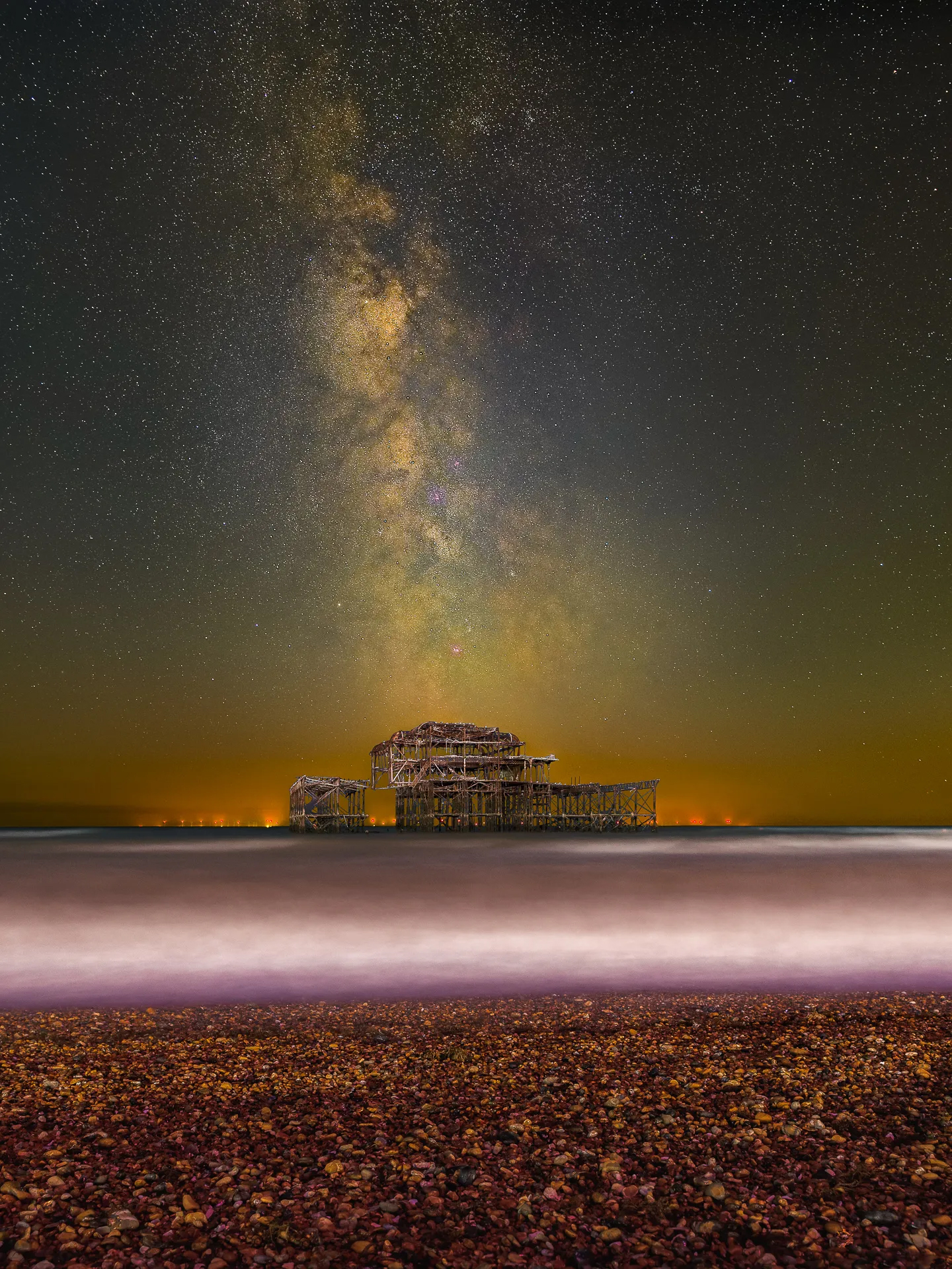 Starry night sky above a skeletal pier, shimmering waves lapping at a pebble-strewn beach, with distant city lights glowing