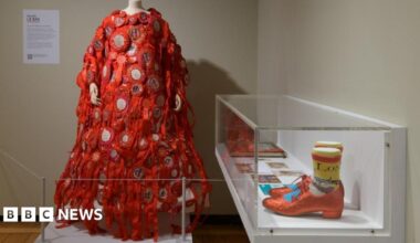 A mannequin displays a red, ribbon-covered dress with circular patches next to a glass case containing glittery red shoes and colourful socks.