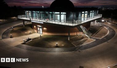 The observatory, seen from outside and from an elevated angle, partly flood lit and partly silhouetted against a sunset