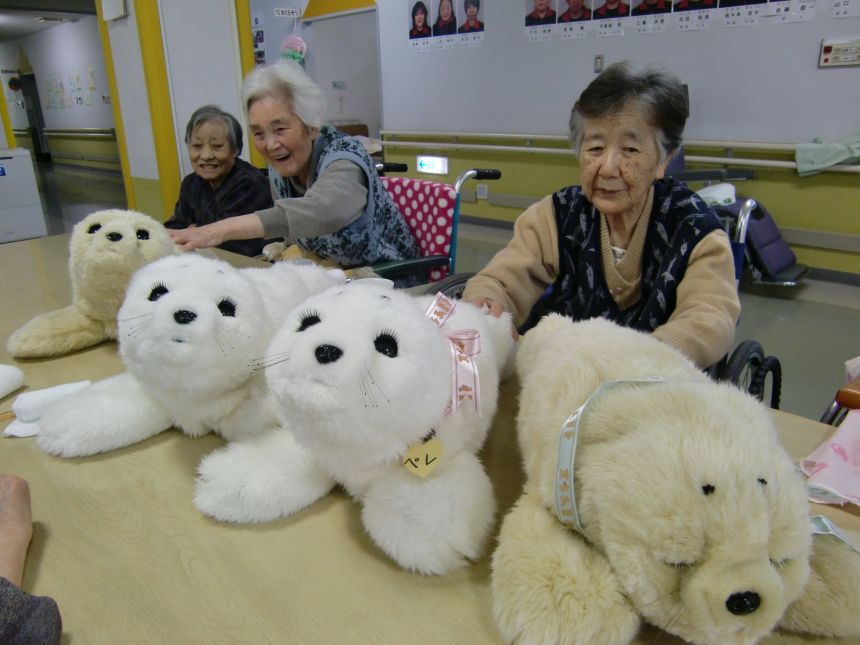 Elderly people using PARO, a therapeutic AI-powered plushie robot companion, designed to look like a baby harp seal. It has been used in Japan since 2005 and throughout Europe since 2023.