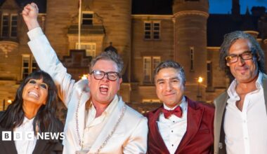 A woman and three men stand shoulder to shoulder in front of a large castle-type building, which is lit by lights at night. To the left, a woman with long black hair, who wears a black jacket and white shirt, smiles as she leans towards a man who is shouting with one arm raised high in the air. He has short light-brown hair and wears a white jacket and shirt. To the right, a man with grey slicked-back hair wears a burgundy dinner jacket, matching bow tie and white shirt, stands next to a man with long black hair, black glasses, a silver jacket and white shirt. They are both smiling.