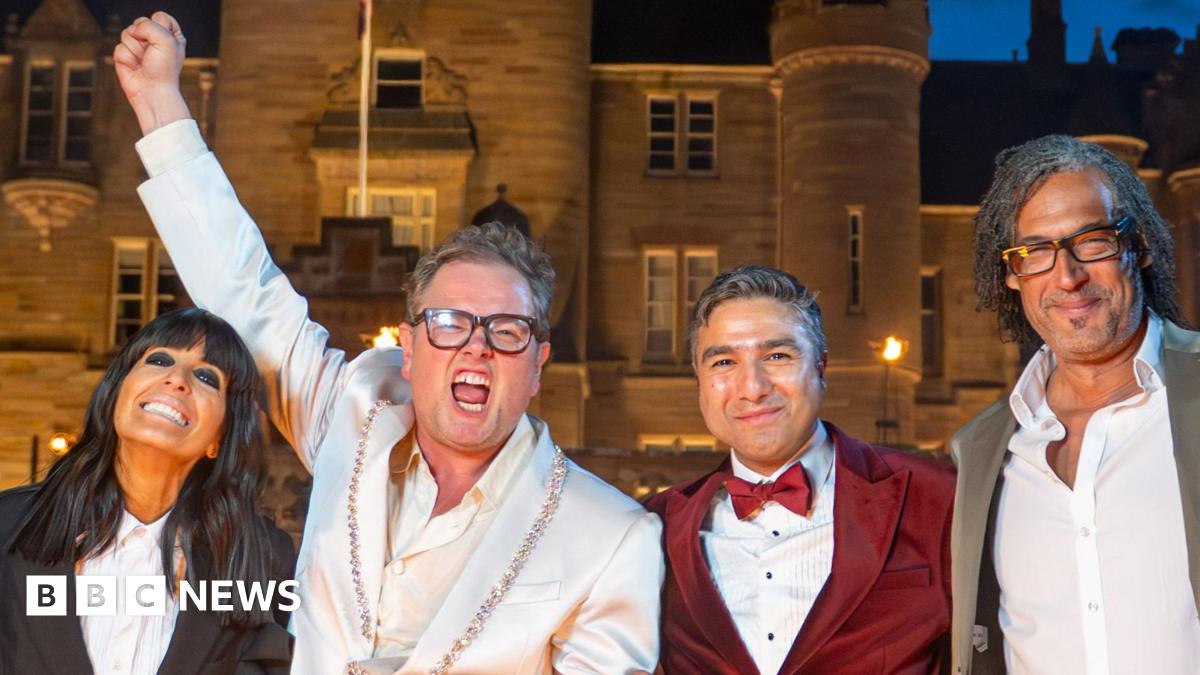 A woman and three men stand shoulder to shoulder in front of a large castle-type building, which is lit by lights at night. To the left, a woman with long black hair, who wears a black jacket and white shirt, smiles as she leans towards a man who is shouting with one arm raised high in the air. He has short light-brown hair and wears a white jacket and shirt. To the right, a man with grey slicked-back hair wears a burgundy dinner jacket, matching bow tie and white shirt, stands next to a man with long black hair, black glasses, a silver jacket and white shirt. They are both smiling.
