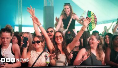 A crowd of people in a big festival tent with hands up and smiling into the camera. Someone is up on their friends shoulders and one girl is holding a "defected records" fan.