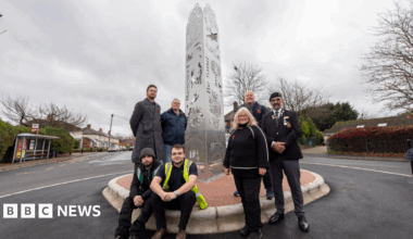 A group of seven people are in front of a metal sculpture, which looks to be in the middle of a roundabout. On the left of the sculpture two of the men are sitting, while two others stand behind them. To the right two men and one woman are all standing