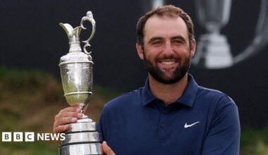 A man in a navy blue polo shirt, white trousers and a silver wrist watch holds up a trophy up and smiles at the camera. Behind him is a putting green and a sign which says "153rd Royal Portrush".