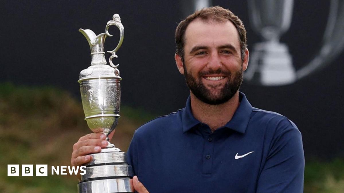 A man in a navy blue polo shirt, white trousers and a silver wrist watch holds up a trophy up and smiles at the camera. Behind him is a putting green and a sign which says "153rd Royal Portrush".