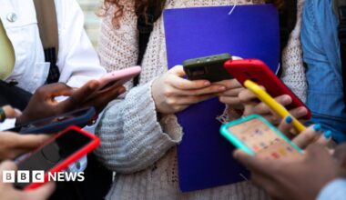 A large number of students stand in a circle on their smart phones in a stock photo. You can only see the hands and blurred phone screens.
