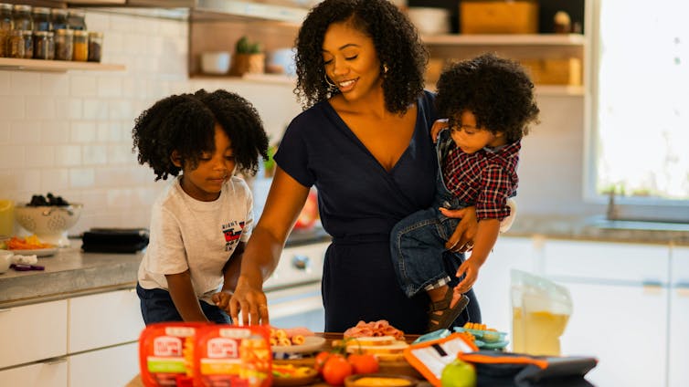 A woman holding a toddler and putting together lunch boxes while another child watches.