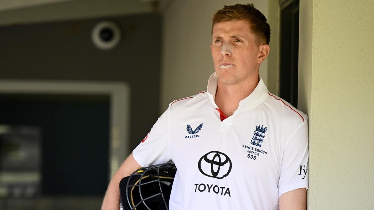 Zak Crawley leans against a wall holding his helmet under his arm