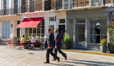 Two businessmen in suits walk past Ottolenghi Deli, while two women sit at an outdoor table under a red awning.