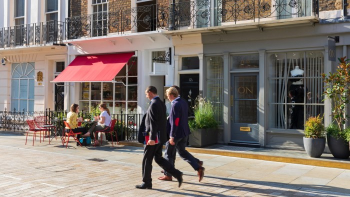 Two businessmen in suits walk past Ottolenghi Deli, while two women sit at an outdoor table under a red awning.