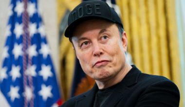 Elon Musk wears a black “DOGE” cap and black jacket, looking to the side, with a blurred US flag in the background.