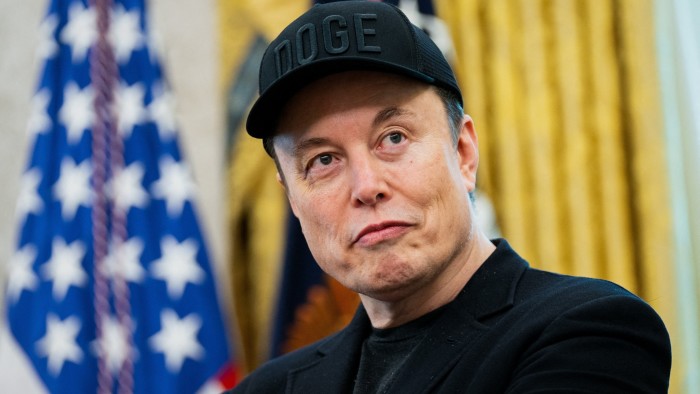 Elon Musk wears a black “DOGE” cap and black jacket, looking to the side, with a blurred US flag in the background.