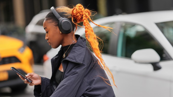 A person with an orange braided bun and ponytail, wearing black headphones and a black cropped jacket, looks at their phone.