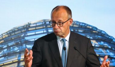 Friedrich Merz gestures while speaking at a podium, with the Reichstag dome visible in the background.