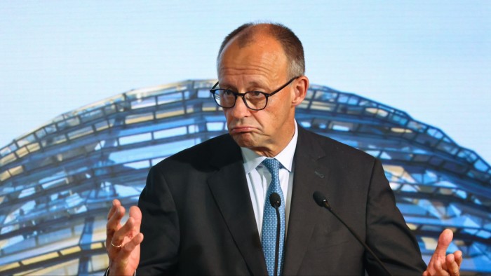 Friedrich Merz gestures while speaking at a podium, with the Reichstag dome visible in the background.