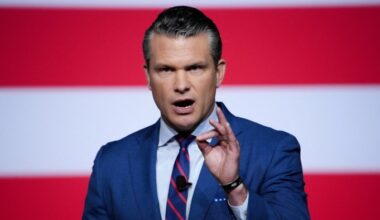 Pete Hegseth speaks with a serious expression, gesturing with his hand, in front of a backdrop with red and white stripes.