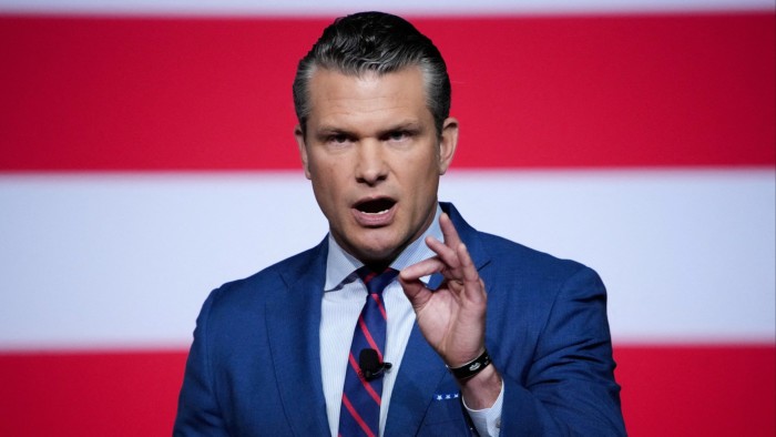 Pete Hegseth speaks with a serious expression, gesturing with his hand, in front of a backdrop with red and white stripes.