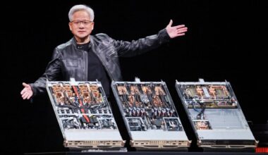 Jensen Huang gestures with open arms while standing behind three open computer server units displaying internal hardware