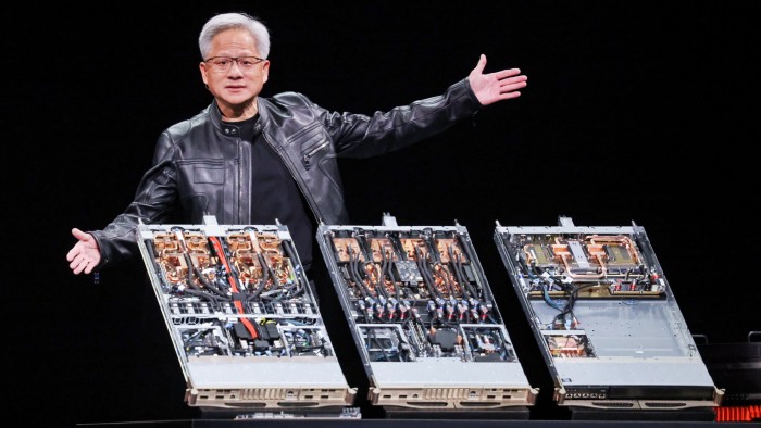 Jensen Huang gestures with open arms while standing behind three open computer server units displaying internal hardware