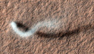 Image of a rough, hilly landscape taken from above. The landscape is barren and reddish. At the left , the white plume of a dust devil stretches upwards before diffusing off to the right.