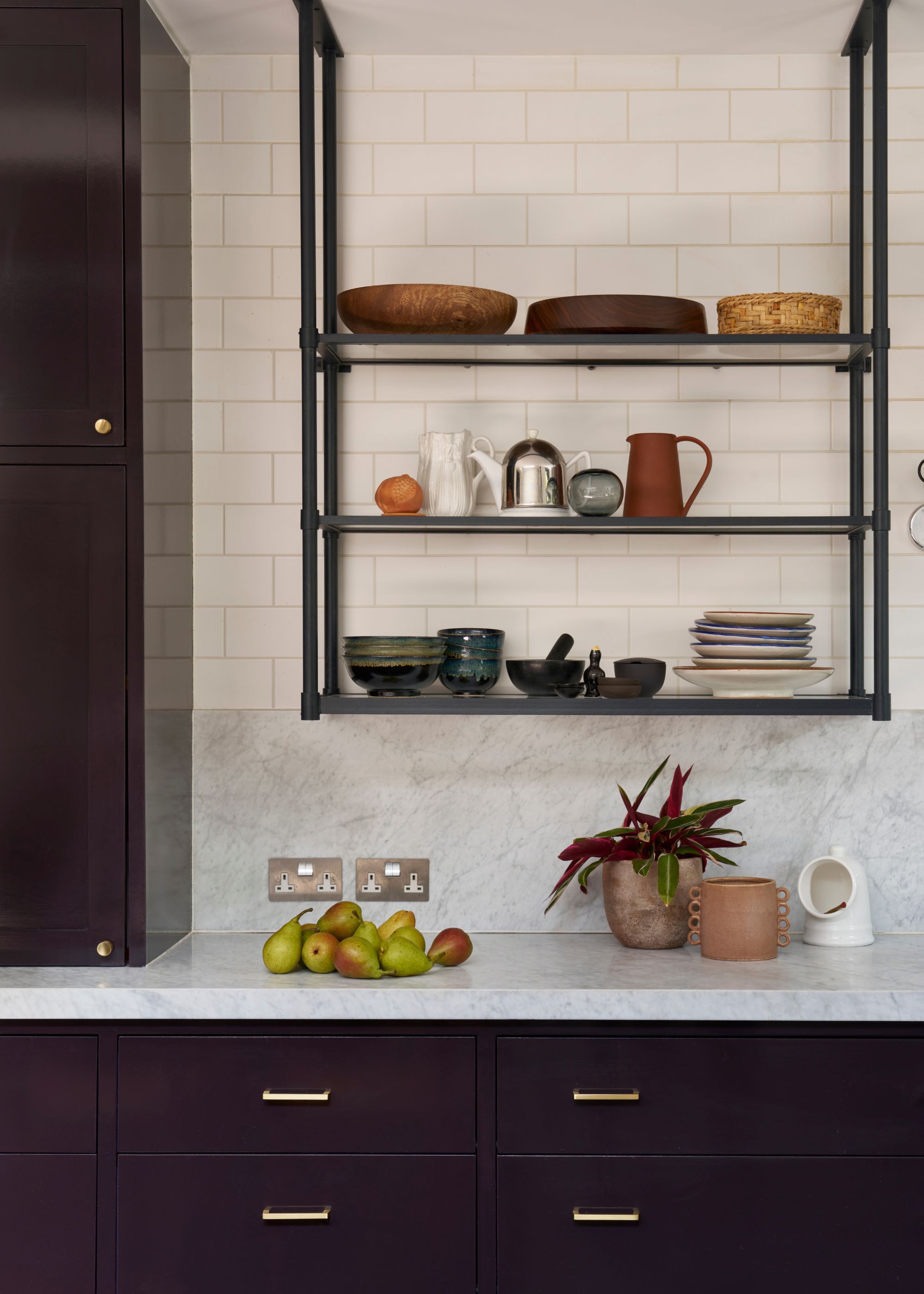 A corner of the kitchen with a view of the kitchen countertop, drawers and open shelving above. On the shelves are dishes and cooking/serving essentials. On the kitchen countertop is a plant pot as well as decor pieces and some a bunch of pears in a pile.