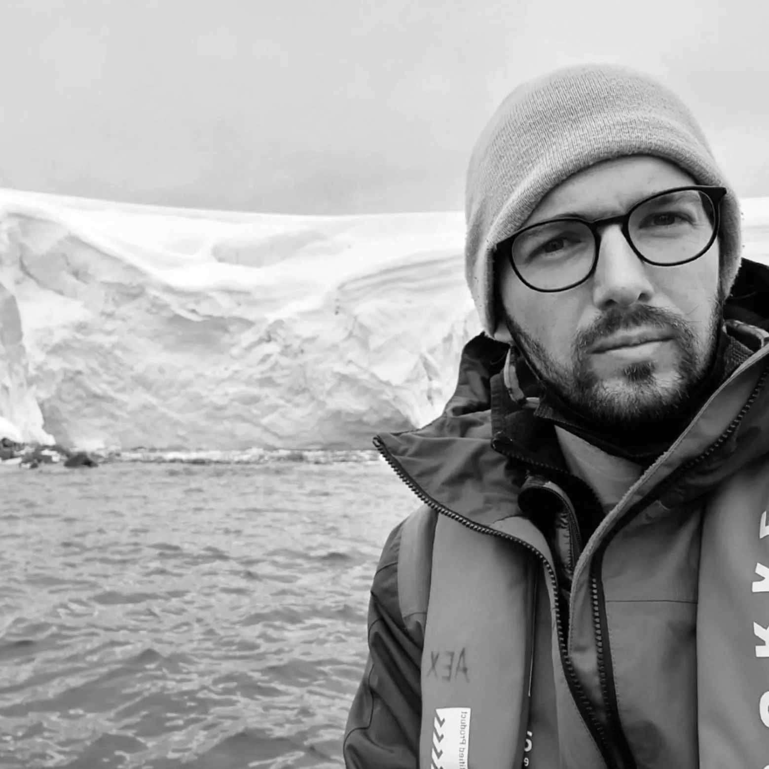 A person in a life jacket stands in front of a massive, icy landscape with snow-capped formations and calm water in a monochrome setting