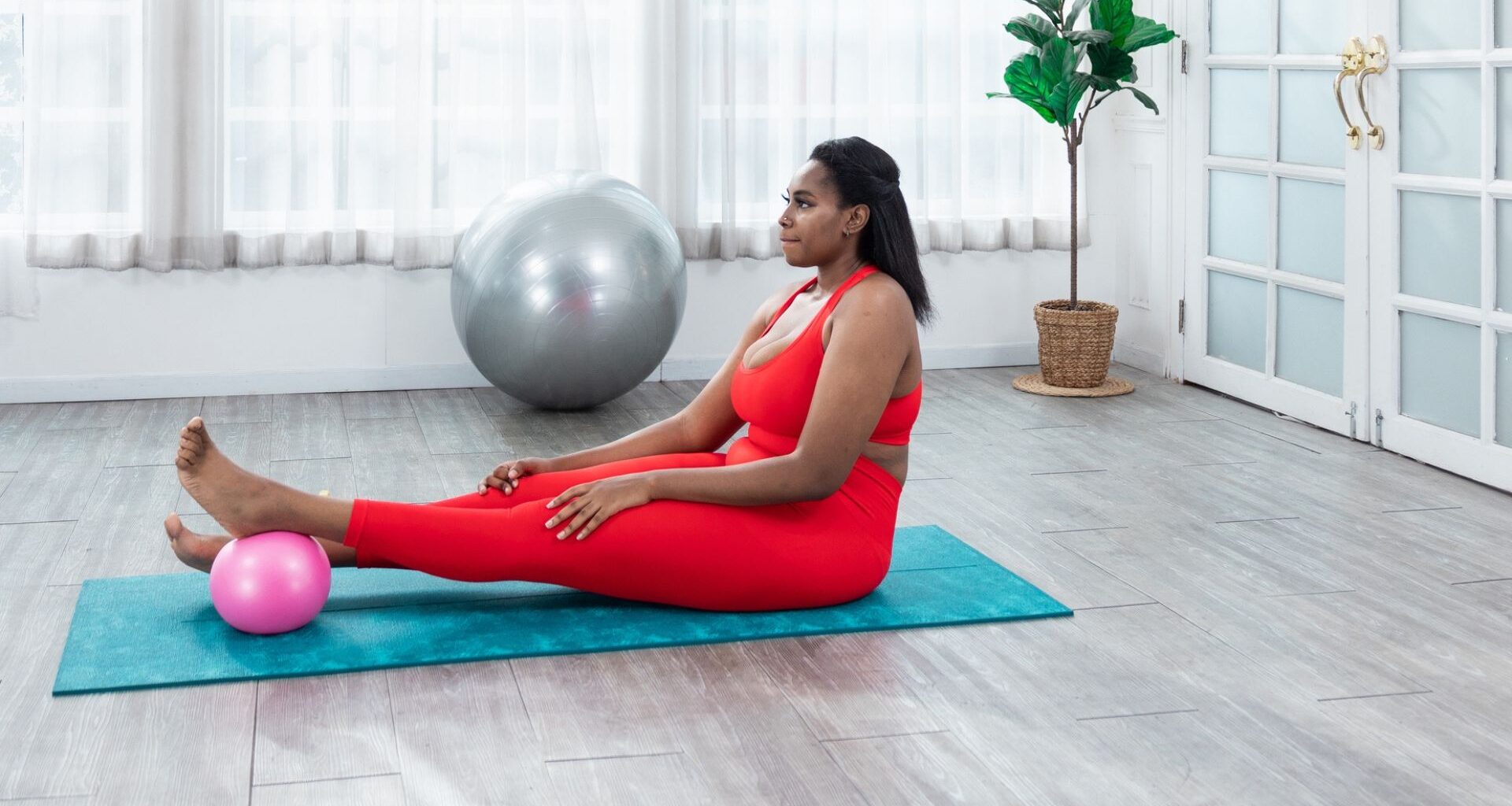 woman wearing red leggings and crop top sitting with legs outstretched on a turquoise exercise mat. there's a pink pilates ball under one foot and she's in a studio setting.