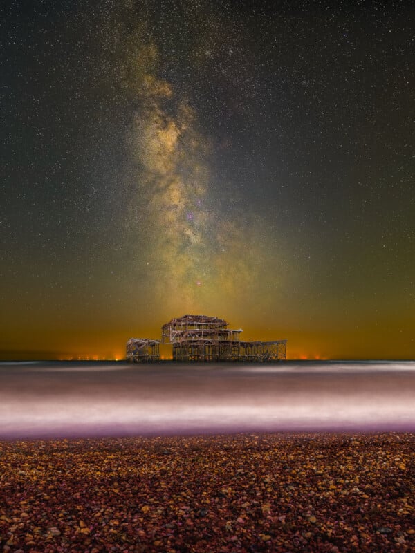 A long-exposure photo of a pebble beach with waves, an old pier’s skeletal remains, and the Milky Way galaxy brightly stretching across a star-filled night sky above the horizon.
