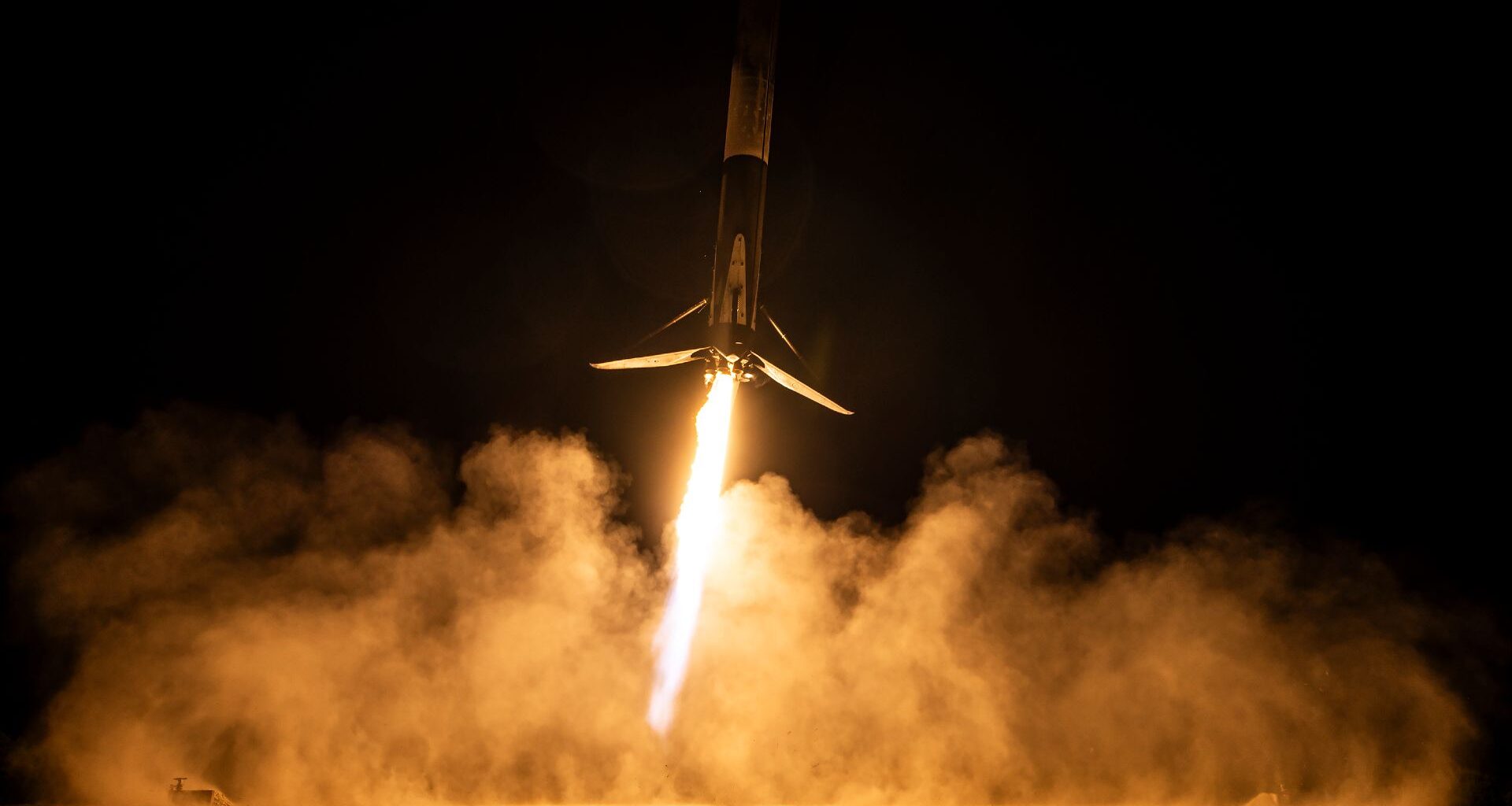 A rocket blazes up into a night time sky with glowing orange clouds of smoke illuminated by the tail of the rocket in the night