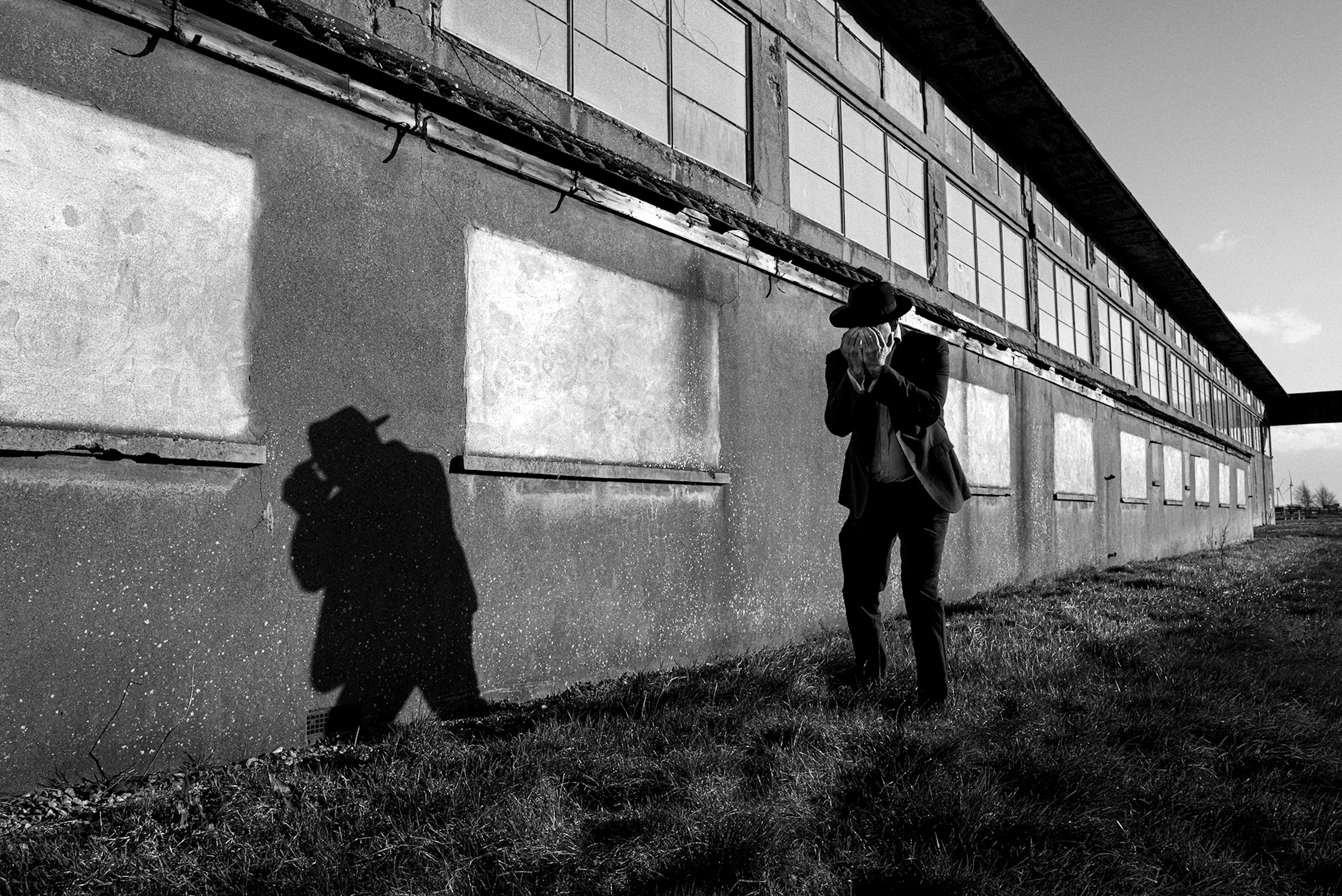 A person in a hat and suit stands against an abandoned building, casting a shadow while holding their head in despair. Black and white image