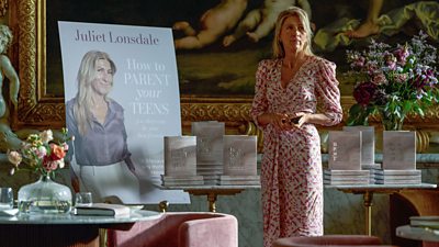 A woman stands in al elaborately decorated room in front of a display of books she's written entitled 'How to Parent Your Teens'. She looks concerned.