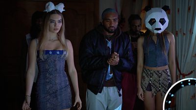 A young woman in a dress and fluffy animal ears, a young man in a dark jacket and grey trousers and another girl in a sleeveless top and skirt wearing a panda mask stand in front of a ring light in a dark room