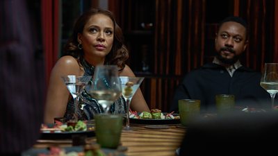 Two people sit at a dinner table looking warily at someone off screen