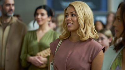 A woman with a curly blonde bob stands among a group of people smiling at something happening out of shot