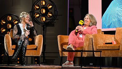 Photo of two women sat in armchairs on a stage. They are both talking into branded BBC microphones. 