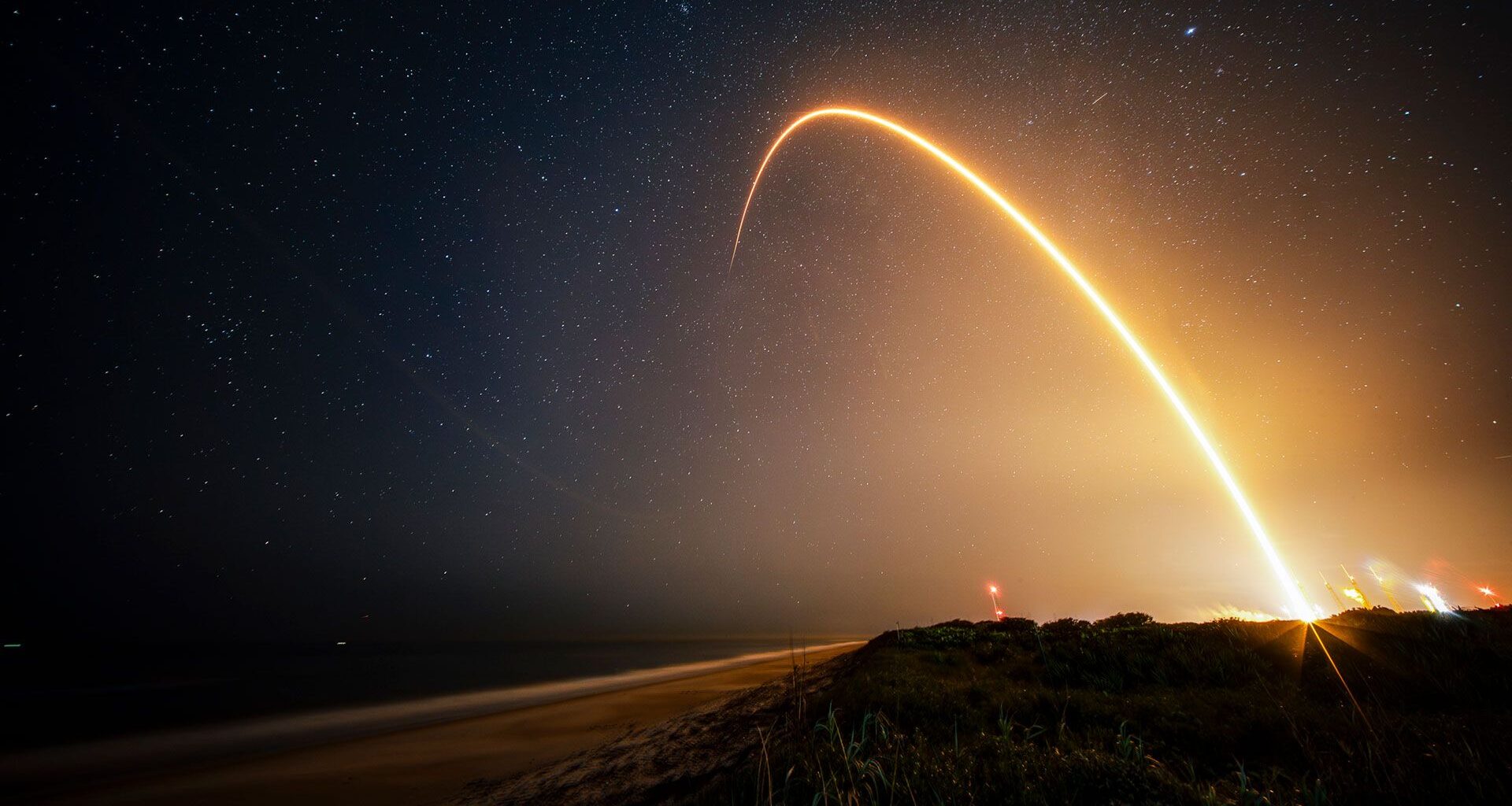 a time-lapse photo showing a streak from a rocket launch arcing over the beach at night.