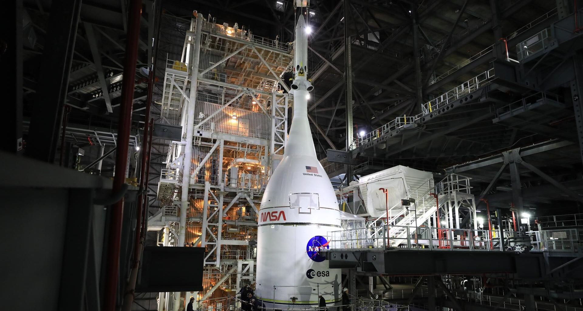 A white pointed rocket module sits on top of a white cylinder, both having the red worm NASA logo and the blue and red meatball NASA logo. The entire system sits in a large warehouse