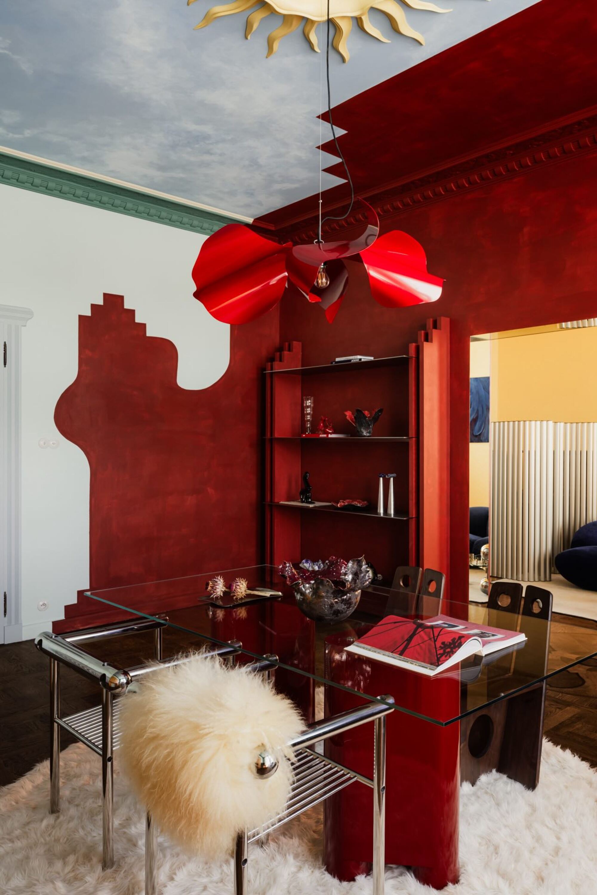 Image of a modern dining room with color-blocked walls in red and light blue. There is a red lacquer table with a glass top that has four metal chairs around it.