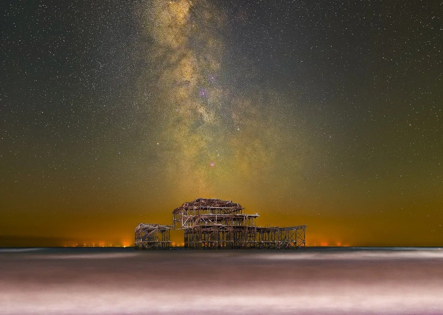 Night photograph of the Milky Way galaxy arcing above the decaying structure of the West Pier in Brighton, with the sea blurred from a long exposure and a pebble beach in the foreground.