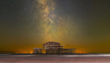 Night photograph of the Milky Way galaxy arcing above the decaying structure of the West Pier in Brighton, with the sea blurred from a long exposure and a pebble beach in the foreground.