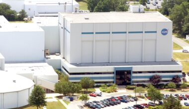 A series of white buildings are seen on a hillside with a parking lot full of cars in the front. One of the buildings has the blue NASA meatball logo on it