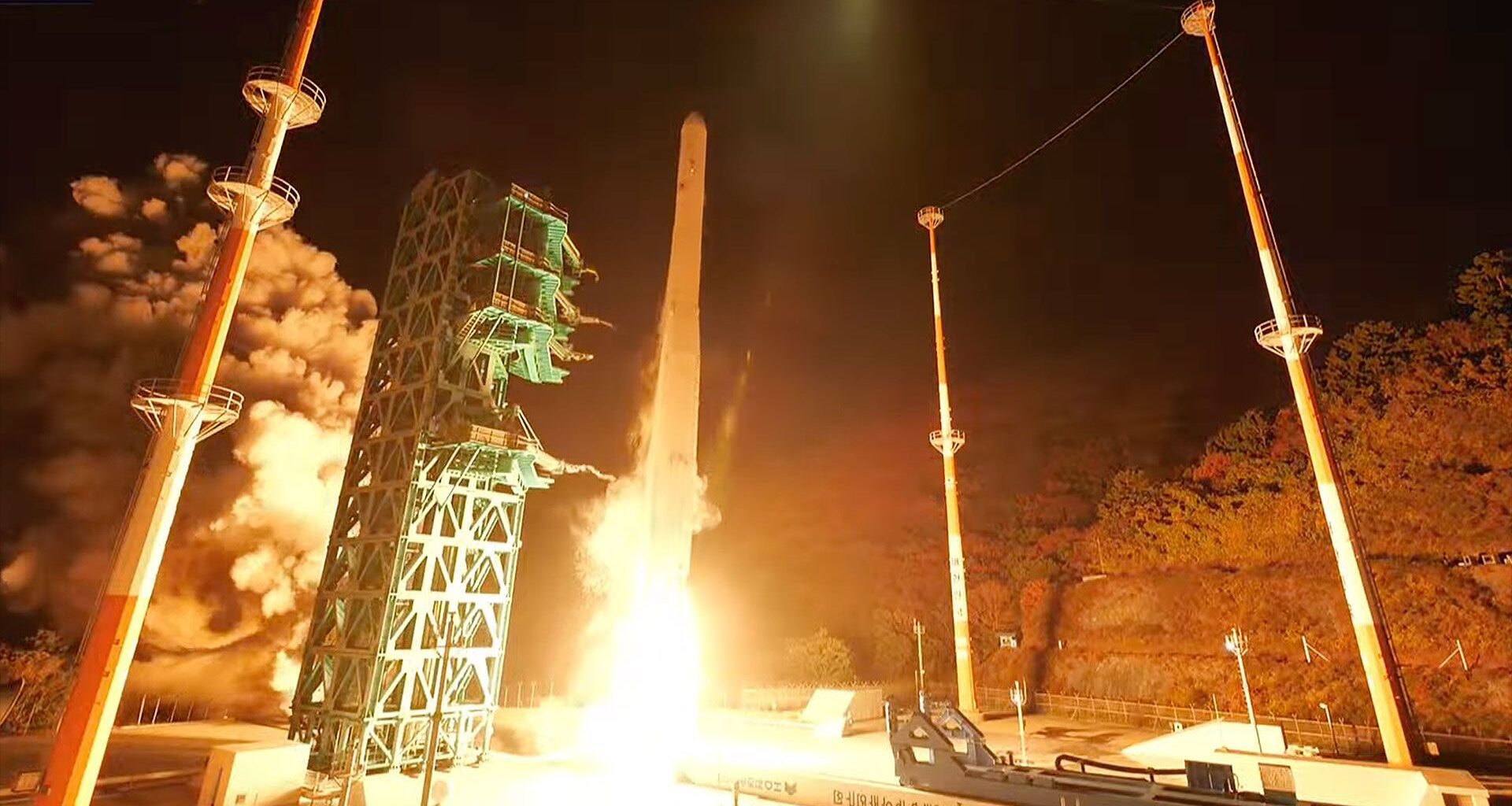 a white rocket lifts off into the night sky, its engine plume lighting the launch pad in white and orange hues.