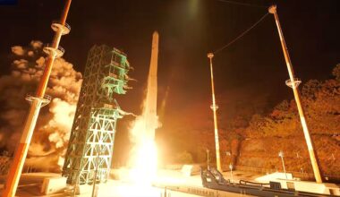 a white rocket lifts off into the night sky, its engine plume lighting the launch pad in white and orange hues.